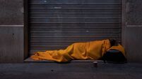 A homeless person sleeps on the street under a blanket in front of a storefront