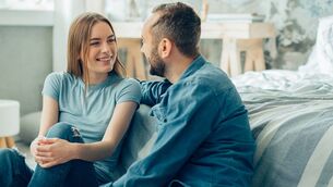 <p>Cheerful young lady smiling to her boyfriend while sitting near the bed next to him Couple talking Couple chatting Picture Stock</p>