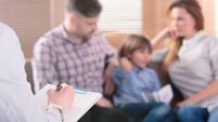 Hand of a professional family psychotherapist writing notes in front of a couple with a child in a blurred background during a c
