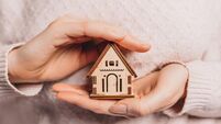 Woman holding a wooden house with her hands with the sun on a light pink background. Sweet home, insurance