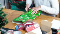 A woman, sitting at a table, wraps a christmas present in brightly coloured christmas wrapping paper. She's trimming the paper t
