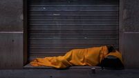 A homeless person sleeps on the street under a blanket in front of a storefront
