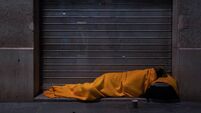 A homeless person sleeps on the street under a blanket in front of a storefront