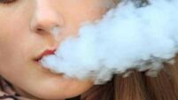 Vape teenager. Young pretty white girl in casual clothing smoking an electronic cigarette opposite modern background on the stre