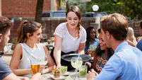 Waitress Serving Drinks To Group Of Friends Sitting At Table In Pub Garden Enjoying Drink Together