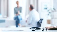 A male senior doctor consults with a female senior patient in a bright, modern office setting. The focus is on a stethoscope and