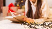 A girl in a white sweater lies and reads a book on a white carpet against the background of a Christmas tree and gifts