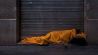 A homeless person sleeps on the street under a blanket in front of a storefront